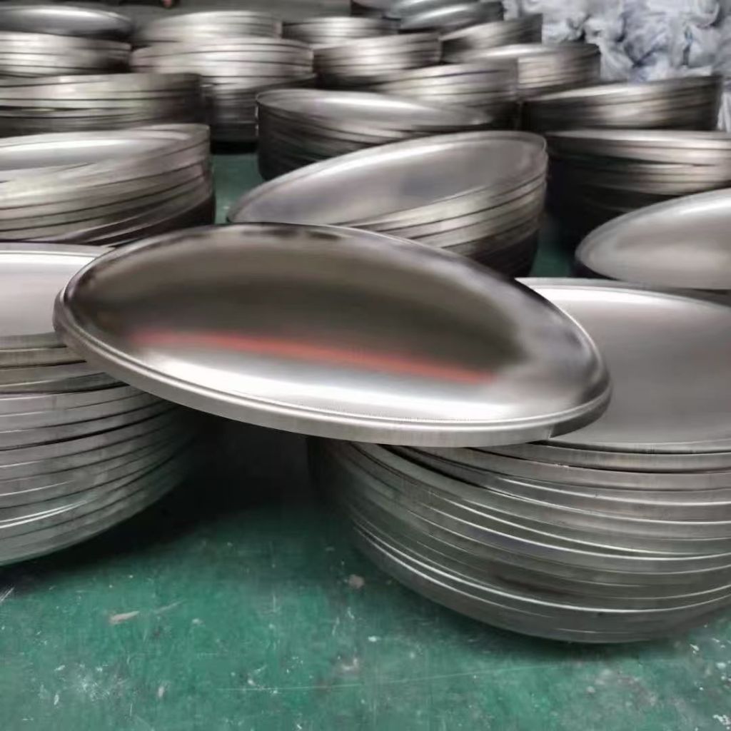Formed stainless steel dished heads with smooth curved profiles stacked in a workshop for tank and pressure vessel fabrication.