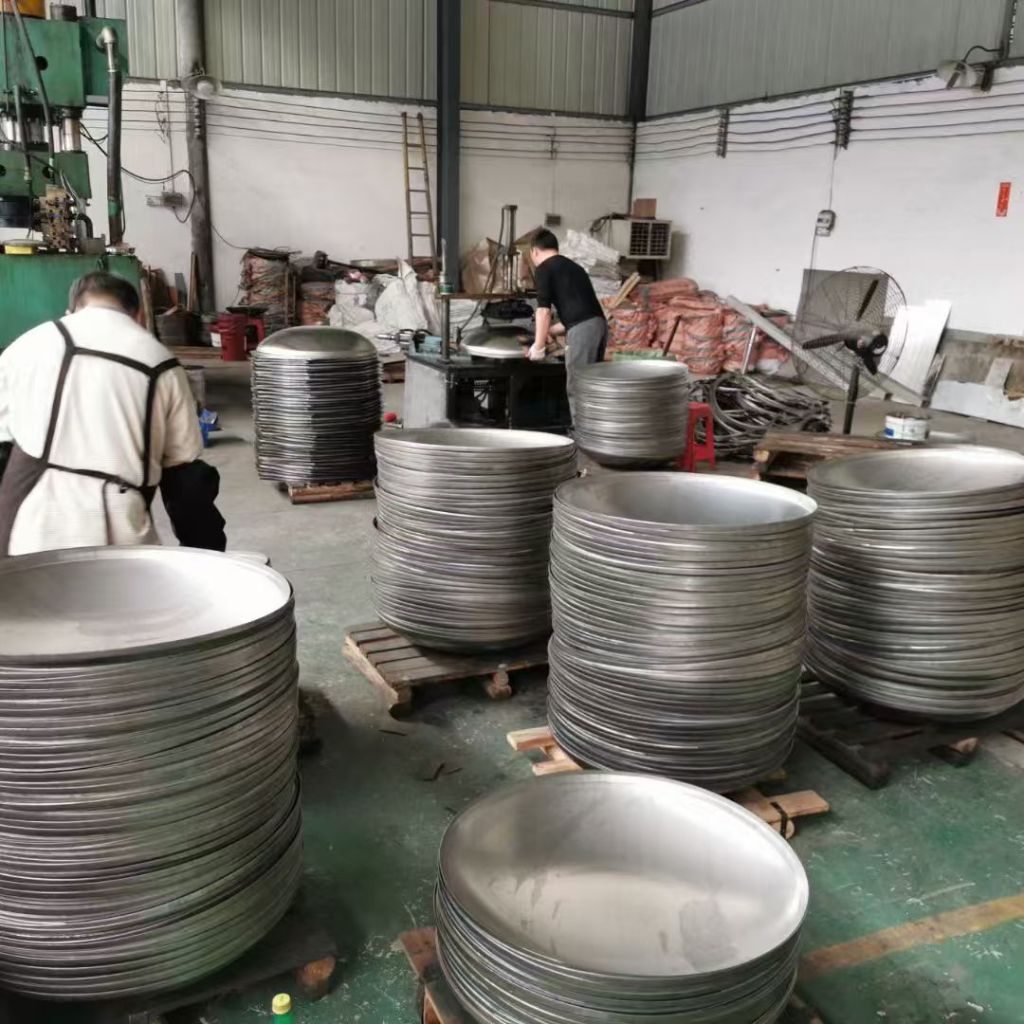 Stacks of stainless steel dished heads in different diameters inside a fabrication workshop for pressure vessel and tank applications.